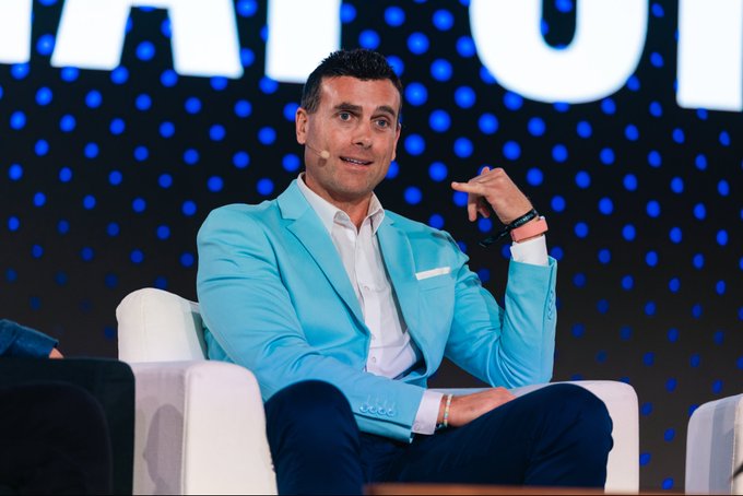 A man in a light blue blazer speaking on stage, seated in a white chair with a dotted blue backdrop behind.