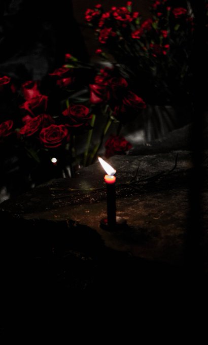 A single lit candle illuminates a bouquet of red roses in the darkness, creating a solemn and intimate atmosphere that reflects the birthday celebrant's quiet, reflective post about turning 30 and minting an OE (Open Edition NFT) to mark the milestone.