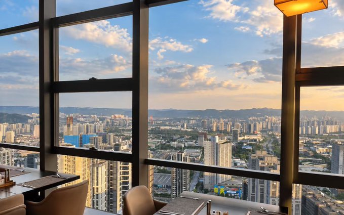 A view of a sprawling city skyline at dusk seen through large floor-to-ceiling windows from a high-rise interior, with a hanging lamp visible.