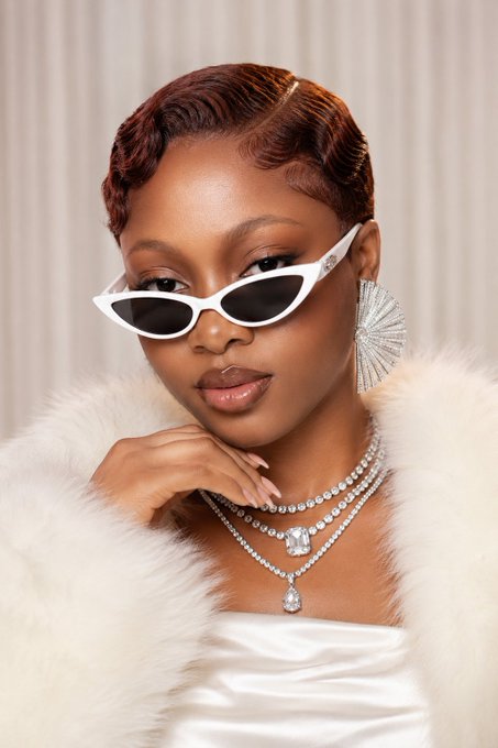A portrait of a woman wearing white cat-eye sunglasses, statement earrings, layered diamond necklaces, and a white fur coat.