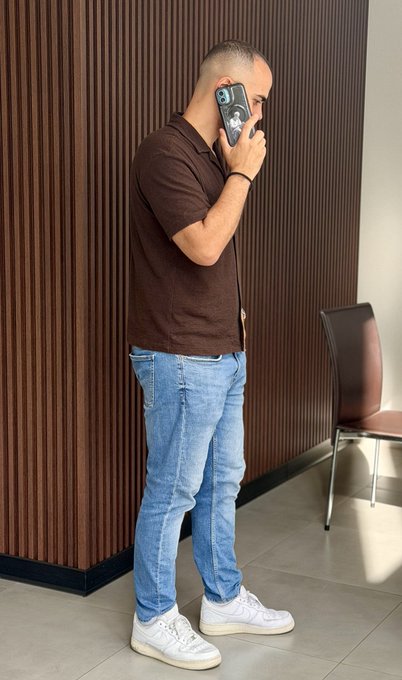 A person taking a mirror selfie while wearing a brown polo shirt, light blue jeans, and white sneakers, standing in front of a wooden slat wall.