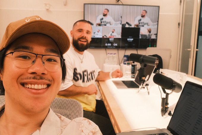 Two men in a podcast studio, one taking a selfie wearing glasses and a cap, the other smiling behind a desk with microphones and a monitor.