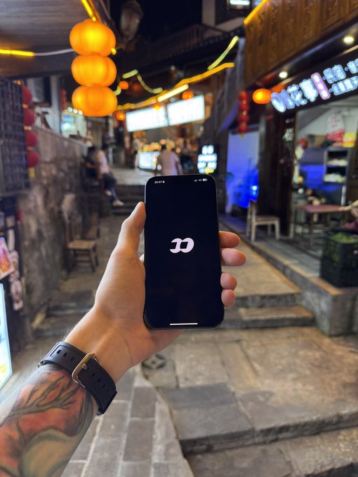 A first-person view of a tattooed arm wearing a black smartwatch, holding an iPhone displaying the iOS boot logo in a street setting with orange Chinese lanterns at night.