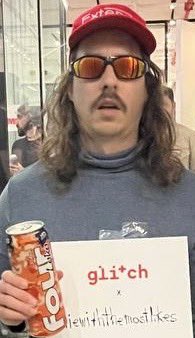 The image features a man wearing sunglasses, holding an orange can of soda, and posing for a picture with a sign that says "gm eth is the ticker. touch grass or buy art you love." The man's sunglasses suggest he might be at an outdoor event or enjoying some time outdoors while drinking his soda.