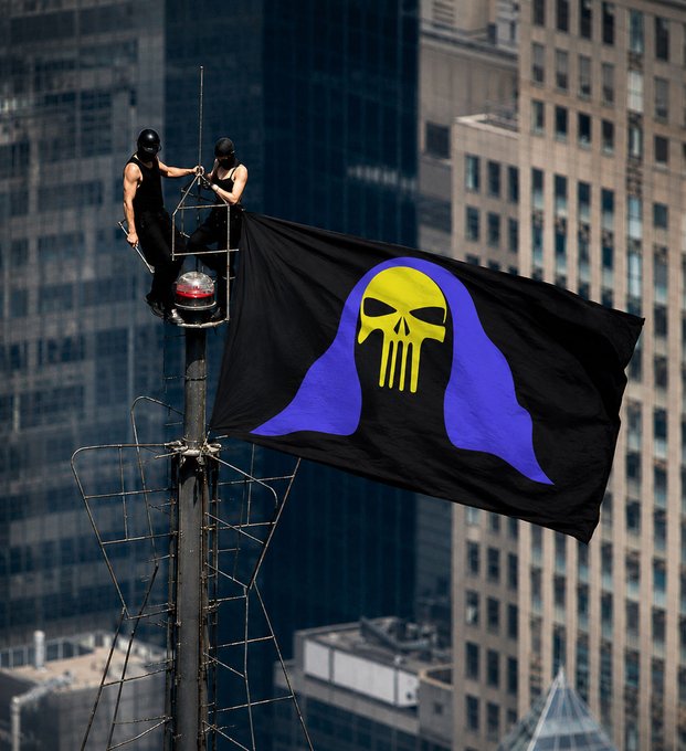 Two people on top of a flagpole fly a large black flag featuring a yellow skull on a blue hooded figure against a city skyline.