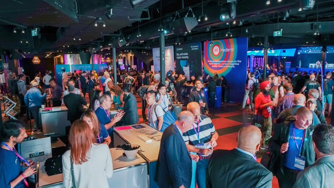 Crowded conference floor with attendees networking under colorful lighting and large screens, one labeled 'Transformative processes take time' with a Network Solutions logo.