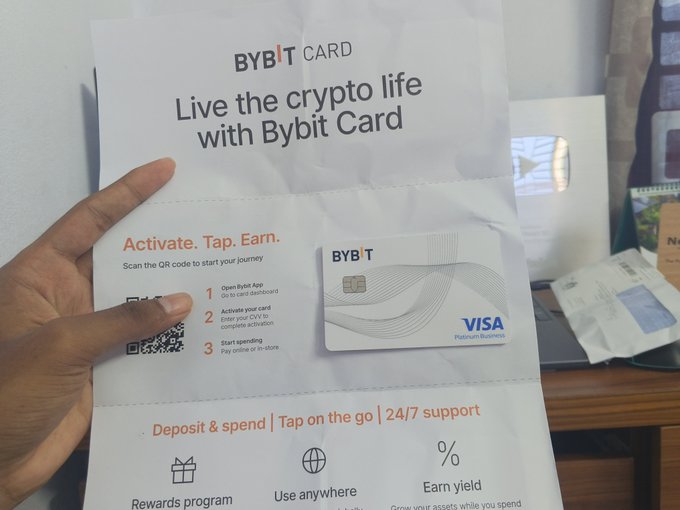 A hand holds a Bybit Card promotional flyer that reads 'Live the crypto life with Bybit Card', featuring a Visa Platinum card and activation instructions.