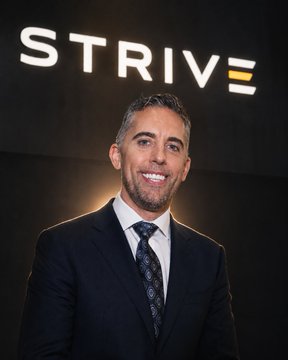 The image depicts a man wearing a suit standing in front of a sign that says "STRIVE" above his head. The background is dark with a spotlight shining directly onto him, highlighting his presence and giving him an air of importance or authority. The sign also mentions that some public companies are buying the company's shares for $2,500 per share.