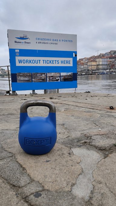 A blue kettlebell with "BOXPT" embossed on it sits on a stone pavement in front of a blue billboard advertising "CRUZEIRO DAS 6 PONTES / 6 BRIDGES CRUISE" and "WORKOUT TICKETS HERE" along the Douro river.