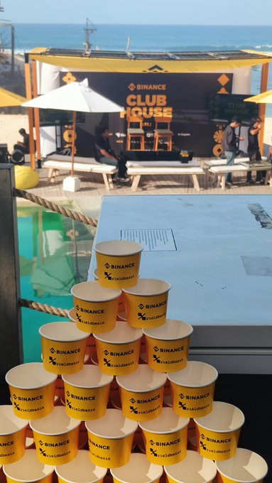 A pyramid of yellow and white 'Binance Clubhouse' cups sits on a table, with a Binance Clubhouse tent and lounge area visible by the beach in the background.