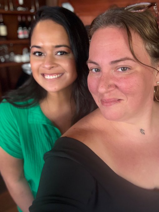 Two women pose for a close-up selfie; one with long dark hair wears a green top and smiles, while the other with sunglasses on her head wears a black off-shoulder top.