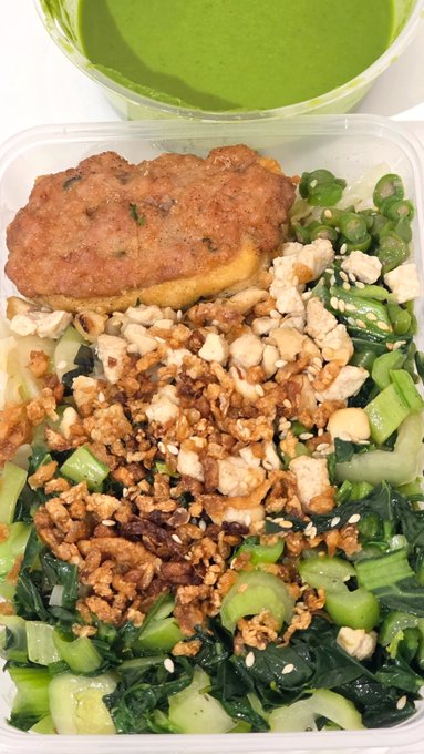 A meal container holds leafy green vegetables, tofu, crumbled topping, sesame seeds, and a breaded item, with a green drink in the background.