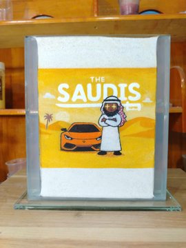 A decorative glass block featuring a 'THE SAUDIS' graphic with a cartoon character wearing traditional Arab clothing and standing next to an orange sports car in a desert scene.
