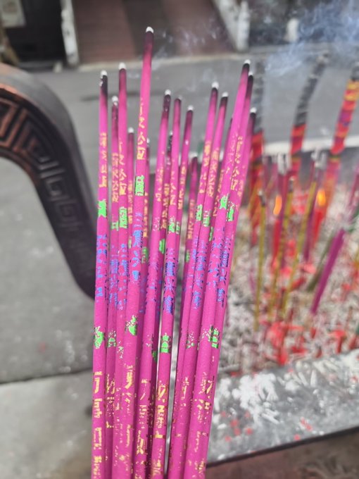 A bundle of pink incense sticks with colorful Chinese characters printed on them, standing upright in a container with incense ash visible at the base.