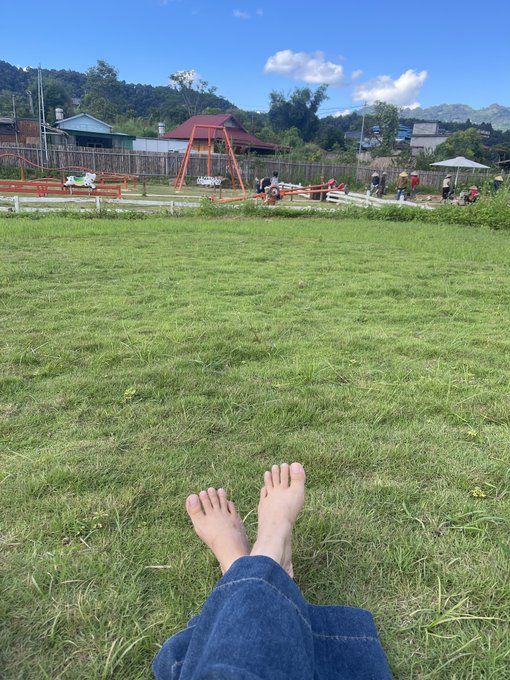A first-person perspective of someone sitting barefoot on grass in a field, looking toward a playground with swings and other people in the distance.