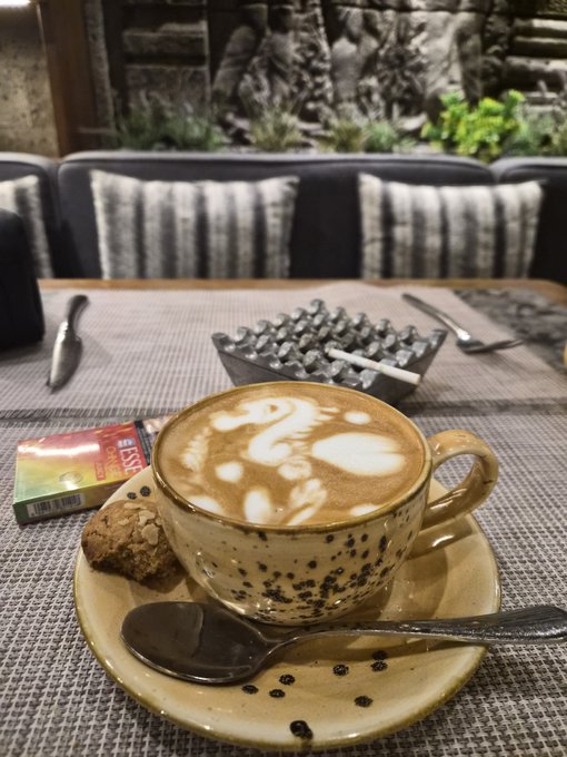 A cozy café setting features a cappuccino with latte art in a speckled ceramic cup, accompanied by an oatmeal cookie and a pack of Davidoff cigarettes, with modern seating in the background. The scene captures the pre-meeting pause suggested by the post, where the coffee and cigarette pack hint at a quiet moment before getting down to business.