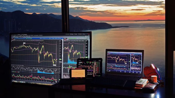 A trading workstation setup with two monitors and a laptop displaying colorful stock charts, set against a stunning ocean sunset view.