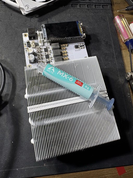 A syringe of Arctic MX-6 thermal paste rests on a CPU heatsink next to a small circuit board and a Duracell battery on a dark workbench.
