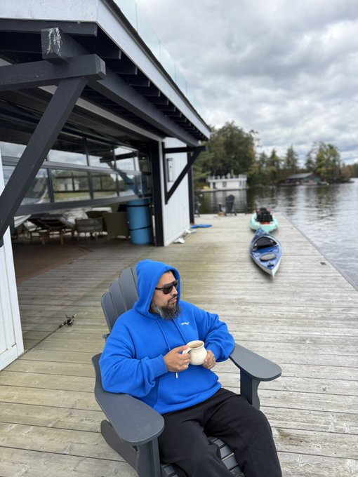 A man in a blue Patagonia hoodie and sunglasses relaxes on an Adirondack-style chair on a lakeside dock, sipping from a mug as kayaks float nearby. The image visually echoes the caption's reflection on the value of different "seats" and quiet moments spent in good company, capturing a peaceful pause by the water with friends.