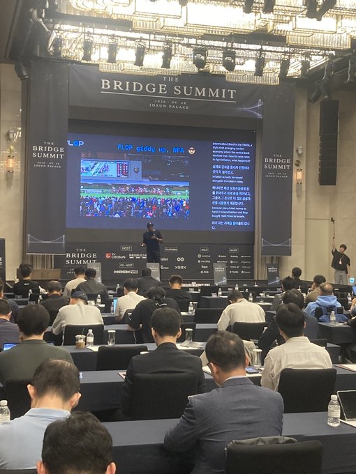 A conference hall audience seated facing a stage with a large screen displaying "THE BRIDGE SUMMIT" logo and "FLOOR giddy up: RIFA" presentation, with various sponsor logos at the bottom.