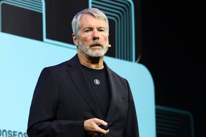 A grey-haired man wearing a dark blazer over a black Bitcoin-branded shirt stands on stage speaking into a headset microphone, with partial text "ONSEOR" visible in the background. The image accompanies the post featuring Michael Saylor's remarks about Bitcoin's evolution into a global digital capital asset, visually representing the speaker delivering this message about future demand drivers like ETFs and corporate balance sheets.