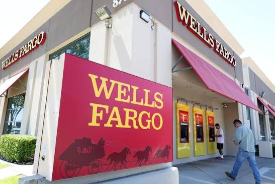 An exterior view of a Wells Fargo bank branch with its distinctive red and yellow signage, showing ATMs and customers outside.