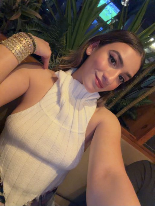 A selfie of a young woman with long brown hair wearing a white ribbed halter top.