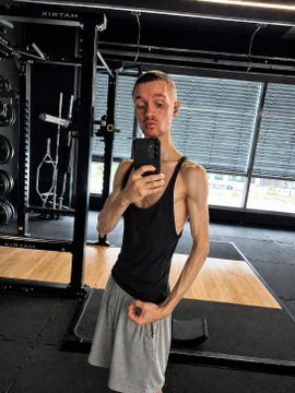 A lean man takes a mirror selfie in a gym wearing a black tank top and gray shorts, with 'KINTAI' visible on the equipment behind him.