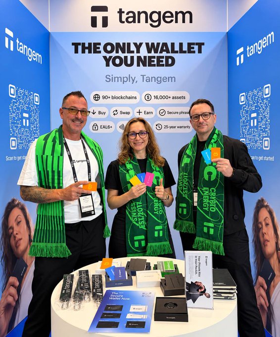 Three Tangem representatives wearing green "Swiss Crypto Valley" scarves pose at IFA Berlin, holding colorful Tangem wallet cards in front of a promotional backdrop reading "THE ONLY WALLET YOU NEED" and "Simply, Tangem," which highlights features like "90+ blockchains," "16,000+ assets," and a "25-year warranty," while a table in the foreground displays Tangem merchandise, product boxes, and promotional materials offering visitors 20% off at Booth 375 in Hall 25.