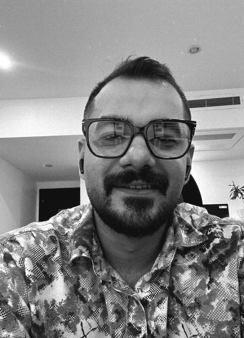 A black and white selfie of a smiling man with a beard, mustache, and glasses, wearing a patterned shirt.