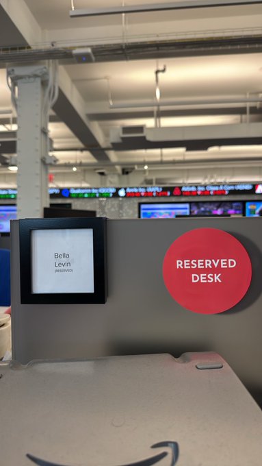 A newsroom office cubicle with a nameplate reading 'Bella Levin' and a large red circular sticker labeled 'RESERVED DESK' on a partition wall.