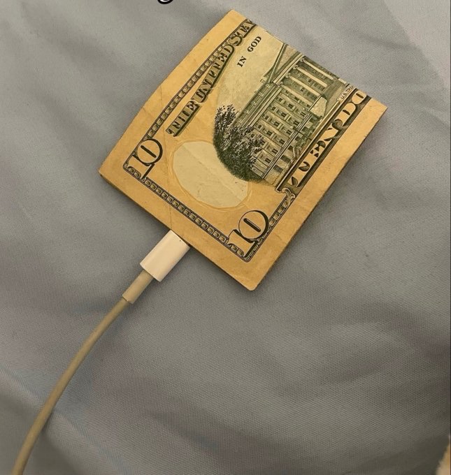 A folded $10 bill is connected to a charging cable like a power bank, lying on a gray fabric surface, symbolizing the irony of trying to "charge" or power up money in hopes of a 100x crypto gain.