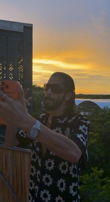 A man with long dark hair, sunglasses, and a black floral shirt taking a selfie with a smartphone against a sunset sky background.