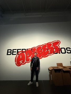A man in a suit and tie standing with arms crossed in front of a large 'BEER BEER'S' logo on a white wall, with cardboard boxes visible to the side.