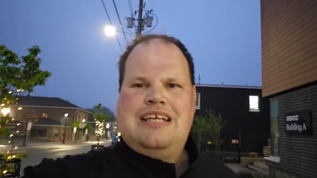 The image features a man standing outside, smiling at the camera with his teeth showing. He appears to be wearing a black jacket or sweater. In the background, there are buildings visible behind him, suggesting an urban setting. The photo also contains text that reads "Me Saying Bitcoin again", which could potentially be related to the Bitcoin statement made by the man in the image.