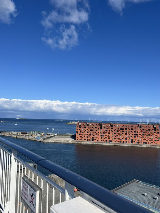 A scenic harbor view from an elevated walkway with a railing, showing calm blue water, a large red brick building, distant ships, and a partly cloudy sky.