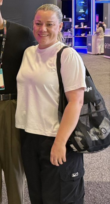 A smiling blonde woman wearing a white t-shirt and carrying a black Slazenger duffel bag stands indoors near neon-lit display shelves.