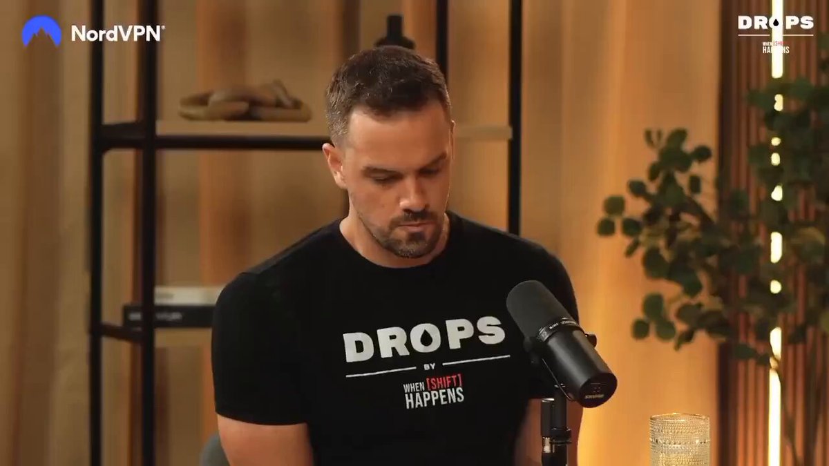 The image shows a man wearing a black t-shirt with white text that says "drops" while sitting at a desk or table, possibly in front of a microphone. The background features a window and a potted plant, creating an indoor setting for the scene.