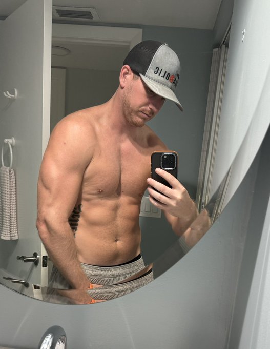 A muscular, shirtless man wearing a gray "Polos" cap and shorts takes a mirror selfie in a bathroom, displaying his abs while flexing one arm. This aligns with his fitness post committing to 150 pushups a day to lose stubborn belly and love handle fat.