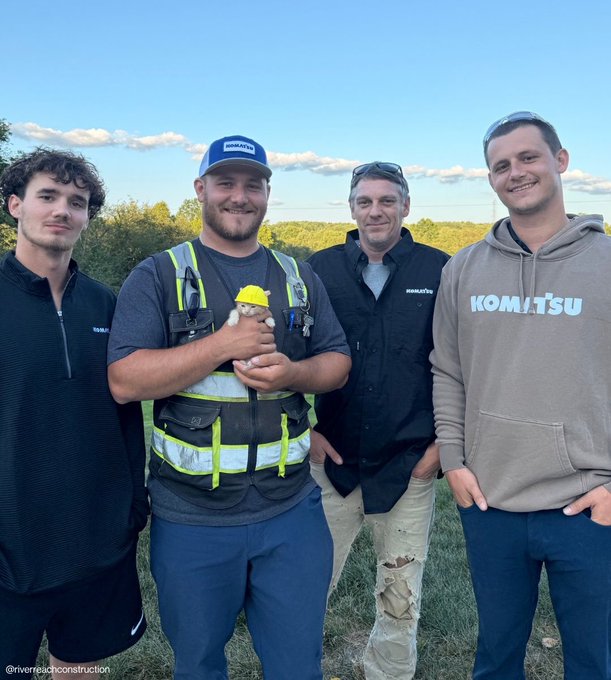 Four men stand together outdoors posing for a photo, with the center man wearing a safety vest and holding a small stuffed toy wearing a yellow hard hat.