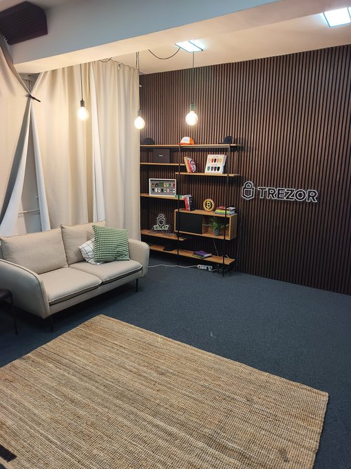 A modern lounge or office space with a sofa, shelving unit, and a wall featuring the 'TREZOR' logo in white lettering on a dark wood-slatted accent wall.