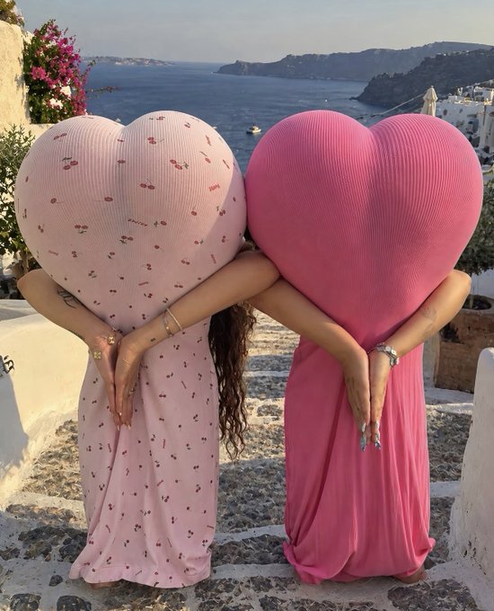 Two women wearing large pink heart-shaped headpieces embrace each other on stone steps overlooking a scenic coastal landscape with the sea and cliffs in the background, suggesting a Santorini-like setting.