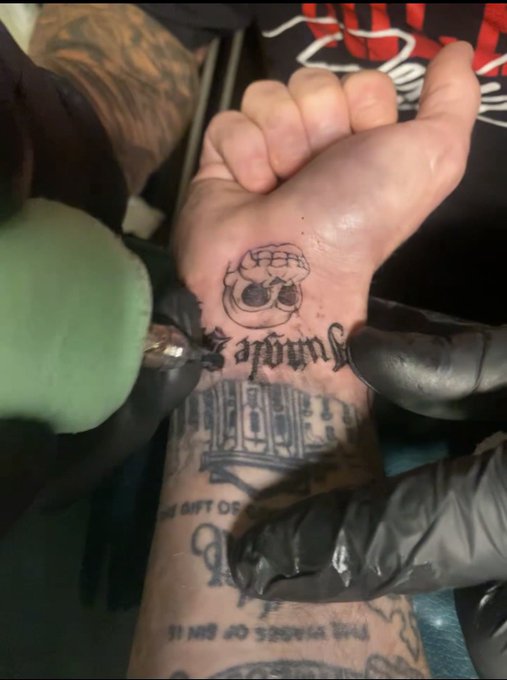 A close-up of a tattoo artist working on a person's foot, adding a small skull tattoo above existing foot tattoos.