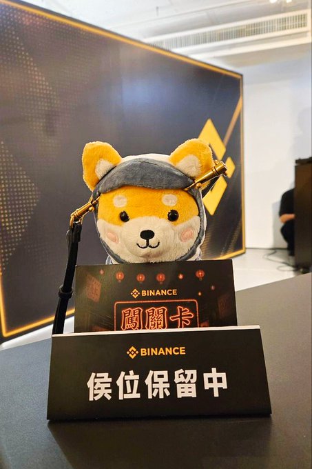A Shiba Inu mascot wearing a grey hood sits behind a Binance-branded "候位保留中" (Seat Reserved) placard and a "關關卡" (checkpoint card) at an offline Binance event, matching the post's description of picking up Binance queue cards while playfully requesting that Binance reserve a spot for the meme coin Asteroid amid tough market conditions and Robinhood's popularity.