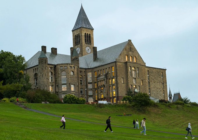 A large stone building with a tall clock tower stands on a grassy hill, with several people walking along a path at dusk.