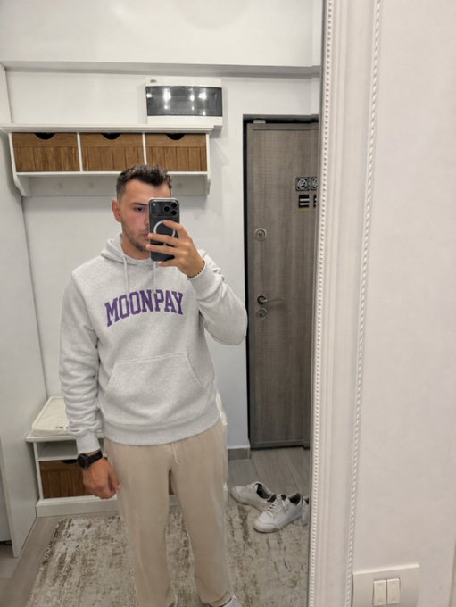 A man taking a mirror selfie wearing a grey 'MOONPAY' hoodie and beige pants, standing in what appears to be an entryway with a door and storage cubbies behind him.