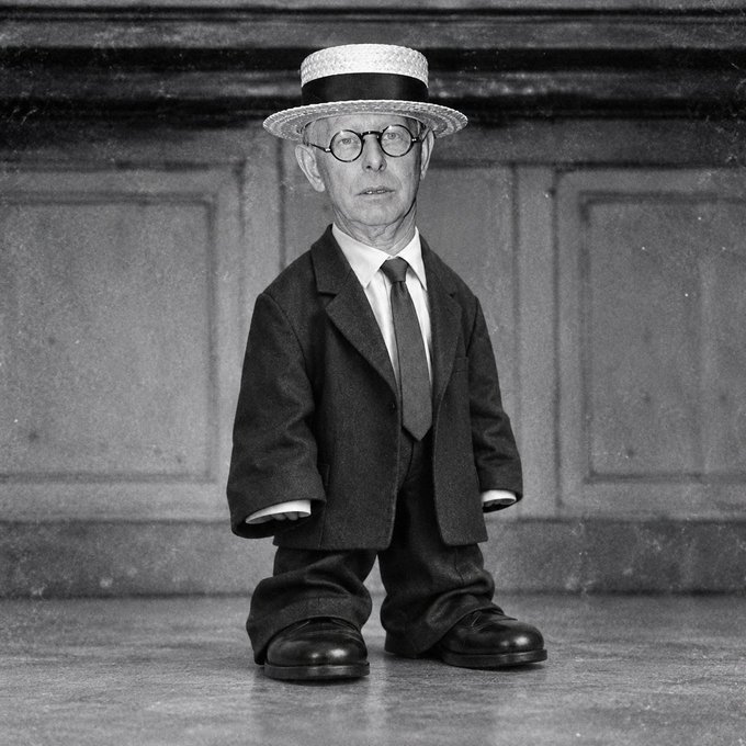 A surreal black and white photograph features a tiny, digitally altered figure of an older man in a suit and straw hat standing against a paneled wall.