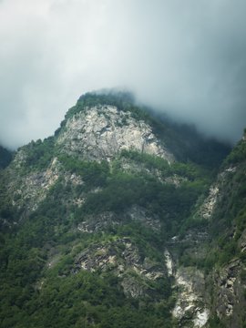iced water cascades down a rocky cliff face, creating a misty backdrop with patches of green vegetation and a gray sky.