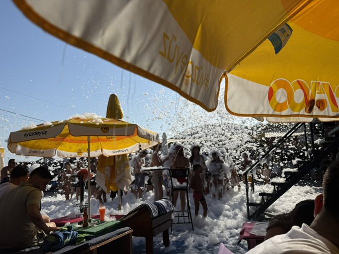 People enjoy a lively foam party under yellow and orange umbrellas branded with "Yandex" and "Yandexy," with the caption humorously noting that "Kurt" showed up to the chaotic, foam-filled beach scene.