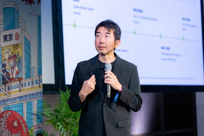 A man in a black blazer speaks into a microphone while presenting in front of a screen displaying a timeline, with a colorful mural featuring 'TOKYO' visible on the left side of the stage.